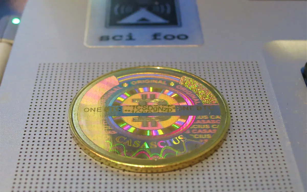 A gold-coloured physical Casascius bitcoin coin with a holographic seal lying on a perforated surface
