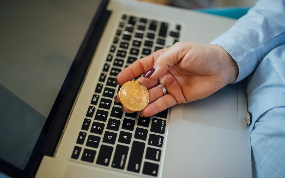 A physical bitcoin coin held in a hand above a laptop (illustrative photo)