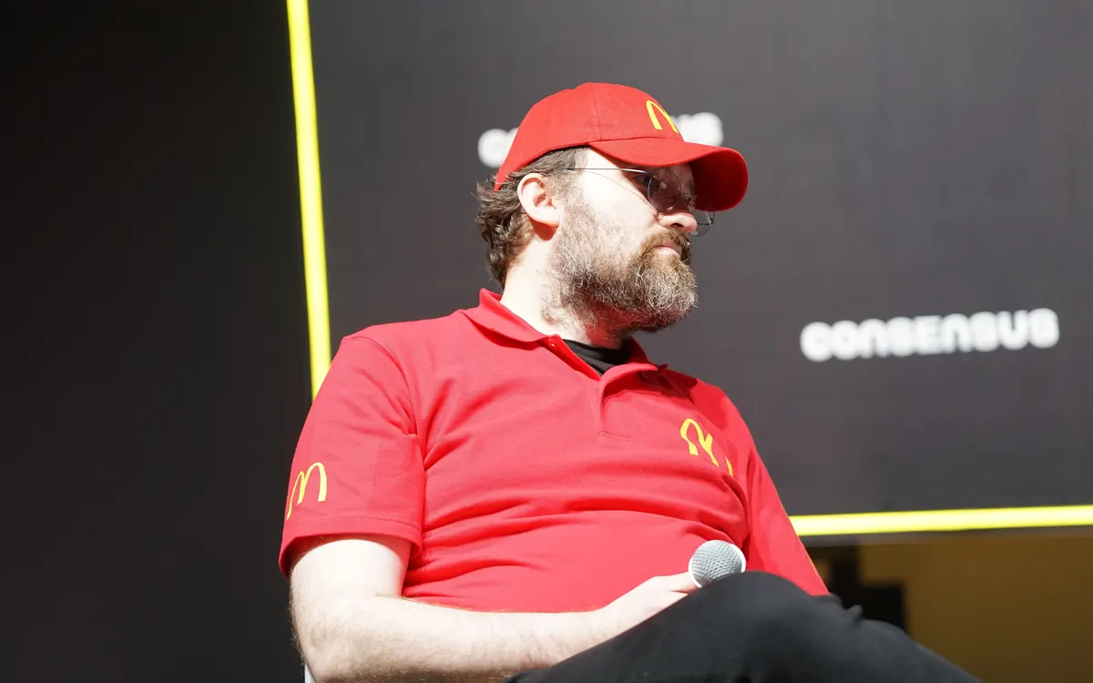 Cardano founder Charles Hoskinson in a red cap and red polo shirt speaking on stage at Consensus 2026