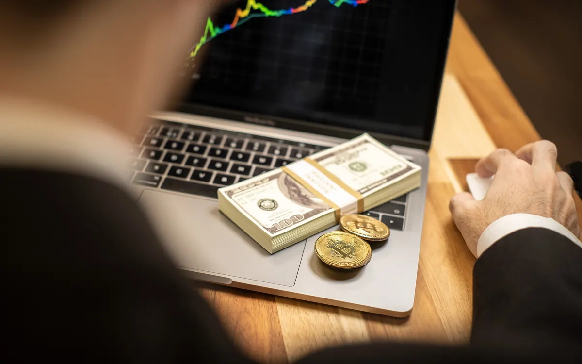 A person at a laptop showing a price chart, with banknotes and gold-coloured crypto coins on the keyboard (illustrative photo)