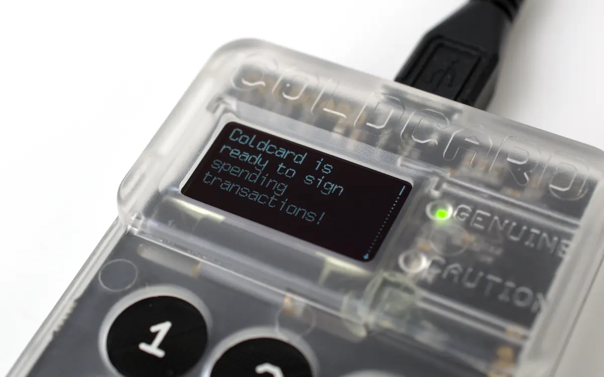 A transparent Coldcard hardware wallet with its screen and a lit 'Genuine' indicator light