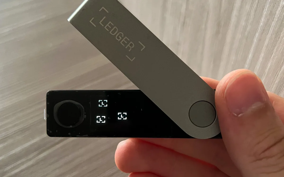 Рука держит аппаратный кошелёк Ledger с небольшим экраном и кнопками (иллюстративное фото)