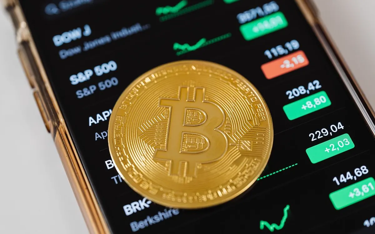 A gold-coloured bitcoin coin resting on a smartphone that shows a list of market prices (illustrative photo)